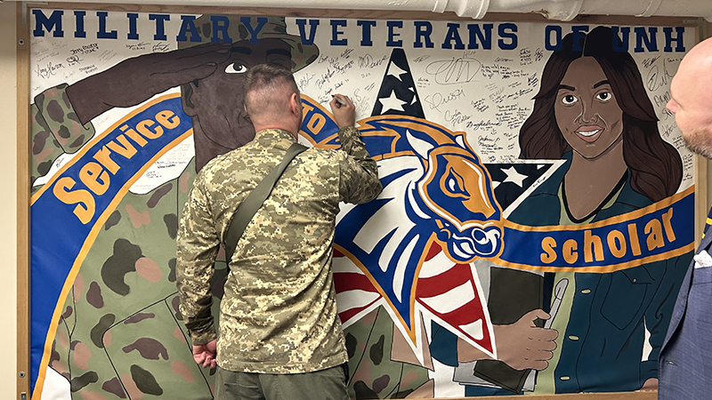 Sr. Lt. Mykola Melnyk signing a wall of honor at University’s Military and Veterans Center, with Ryan Noonan, Manager of Military & Veterans Affairs looking on.