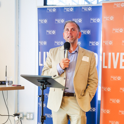 Greg Overend accepted the 'Live United' award on behalf of the University of New Haven. He holds a microphone and stands in front of a red and blue step and repeat